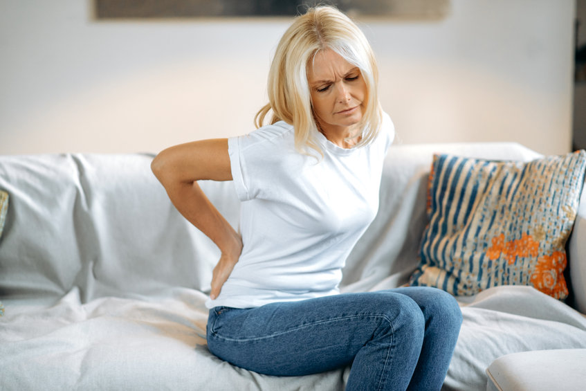 Upset frustrated mature woman suffering from backache, unhappy senior blonde sitting on a sofa at living room, feeling discomfort because of pain in back, have chronic joint pain problems, need rest